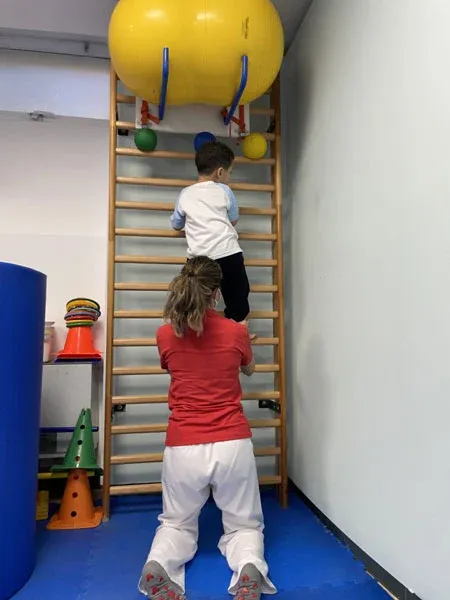 Niño subiendo por las espalderas con ayuda de su fisioterapeuta en atención temprana